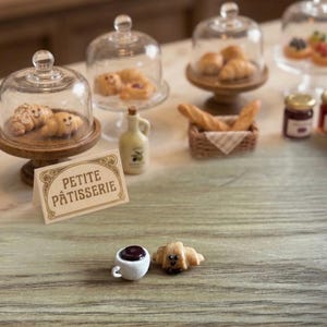 May include: A miniature bakery set featuring glass cloches on wooden stands containing tiny pastries, a small woven basket with bread, a ceramic bottle, a sign reading PETITE PÂTISSERIE, and a small white coffee cup next to a tiny croissant.