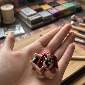 May include: A small, handcrafted clay figurine of a brown and white puppy with a pink bow on its head, nestled inside a cream-colored, textured container. The puppy has dark brown ears, a black nose, and white accents around its eyes and muzzle.