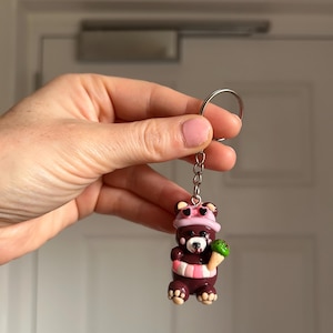 May include: A dark brown bear keychain featuring a light pink hat with heart-shaped accents, a pink and white striped scarf, and a small green ice cream cone held in one paw. The bear has light tan paw pads and a silver metal keyring attachment.