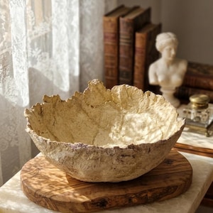Puede incluir: Un cuenco decorativo beige con una superficie texturizada y un borde irregular. El cuenco se apoya en una tabla de madera, colocada sobre una superficie de mármol blanco. El fondo incluye libros antiguos y un busto.