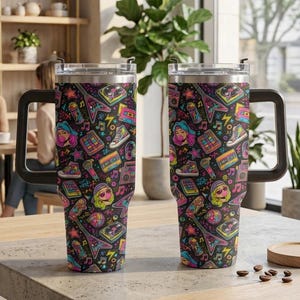 May include: Two stainless steel tumblers with handles featuring a vibrant 1980s-inspired pattern on a black background, including illustrations of cassette tapes, boomboxes, high-top sneakers, electric guitars, and colorful geometric shapes.