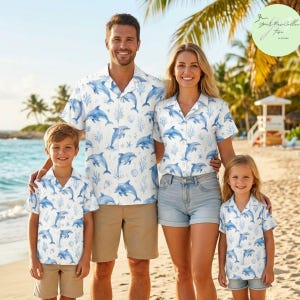 May include: A family wearing matching white short-sleeved button-down shirts featuring a blue dolphin and coral pattern. The shirts are paired with khaki and light blue denim shorts, creating a coordinated look for a beach setting.