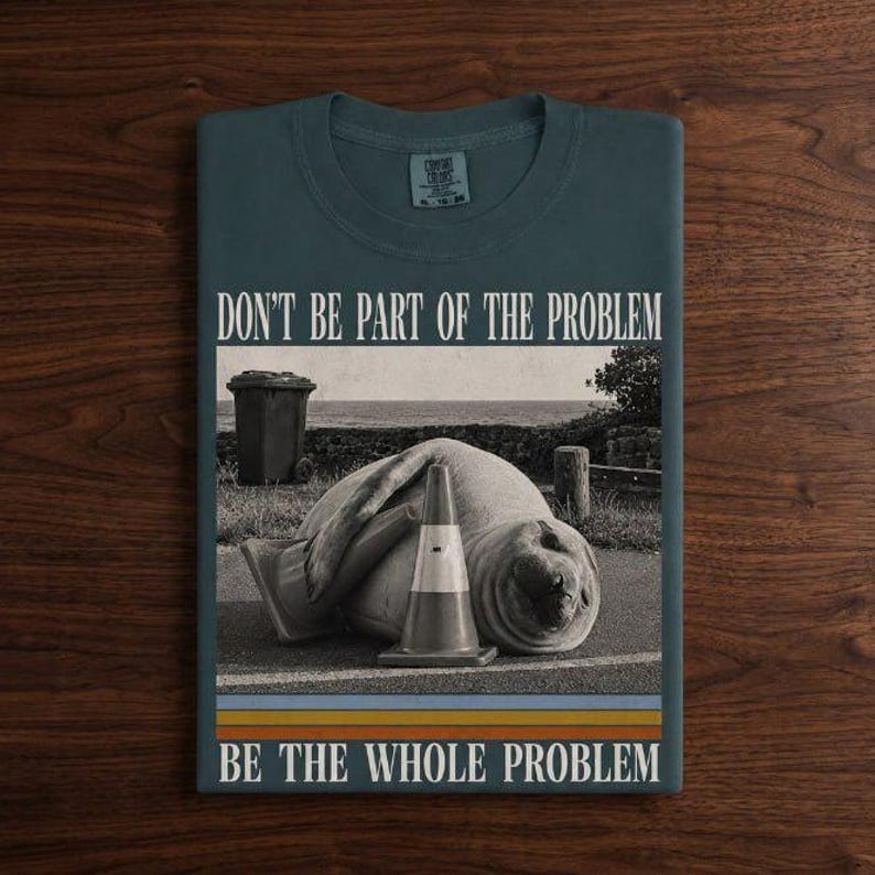 May include: A dark teal t-shirt featuring a graphic of a seal lying next to a traffic cone and a trash can. The text reads "DON'T BE PART OF THE PROBLEM" at the top and "BE THE WHOLE PROBLEM" at the bottom, with horizontal stripes below the image.