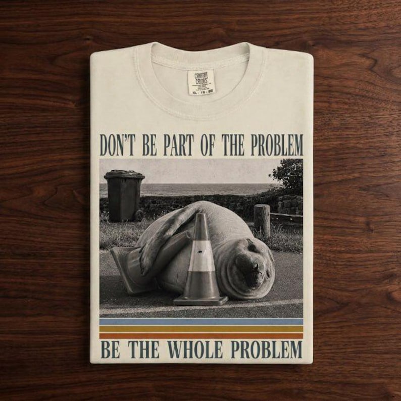 May include: A cream-colored t-shirt featuring a black and white graphic of a seal lying next to a traffic cone. The text reads DON'T BE PART OF THE PROBLEM at the top and BE THE WHOLE PROBLEM at the bottom, with horizontal stripes below the image.
