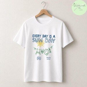 Peut inclure: Un t-shirt blanc &agrave; manches courtes avec un motif comprenant le texte 'EVERY DAY IS A SUN DAY', un soleil jaune et une corde &agrave; linge avec un bikini, un chapeau et des lunettes. Sous le motif, le texte indique 'WITH A CHANCE OF SUN' et 'MOSTLY SUNNY'.