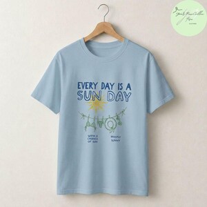 Peut inclure: Un t-shirt bleu clair avec un motif repr&eacute;sentant un soleil jaune et une corde &agrave; linge avec des maillots de bain et un chapeau. Le texte indique : EVERY DAY IS A SUN DAY, WITH A CHANCE OF SUN, et MOSTLY SUNNY. Le t-shirt est sur un cintre.