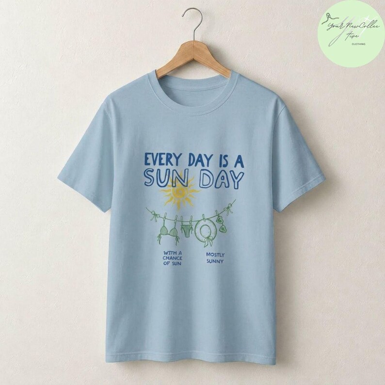 Peut inclure: Un t-shirt bleu clair avec un motif repr&eacute;sentant un soleil jaune et une corde &agrave; linge avec des maillots de bain et un chapeau. Le texte indique : EVERY DAY IS A SUN DAY, WITH A CHANCE OF SUN, et MOSTLY SUNNY. Le t-shirt est sur un cintre.