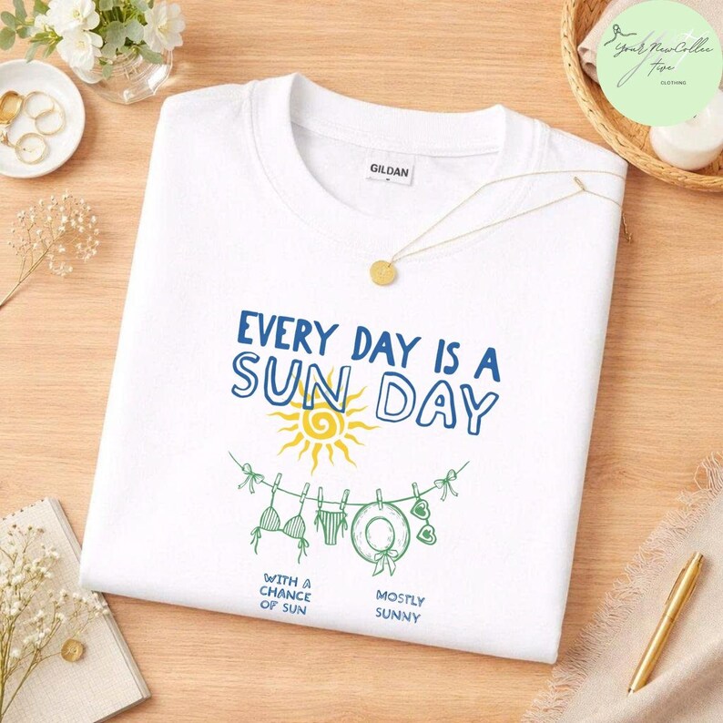 Peut inclure: Un t-shirt blanc &agrave; manches courtes avec un motif bleu et jaune portant le texte "EVERY DAY IS A SUN DAY" au-dessus d'un soleil jaune et d'une corde &agrave; linge verte avec un bikini, un chapeau et des lunettes, et le texte "WITH A CHANCE OF SUN" et "MOSTLY SUNNY".