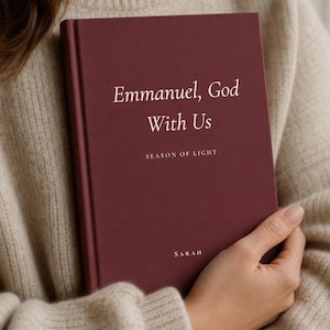 Peut inclure: Un livre à couverture rigide bordeaux profond avec du texte blanc sur la couverture indiquant Emmanuel, God With Us, Season of Light, et le nom Sarah en bas. Le livre a une finition mate lisse et est tenu contre un pull en tricot beige.