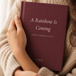 Peut inclure: Un livre à couverture rigide bordeaux profond avec du texte blanc sur la couverture indiquant A Rainbow Is Coming, HOPE CARRIED AGAIN, et le nom SARAH en bas. Le livre est tenu contre un pull en tricot beige, présentant une finition mate.