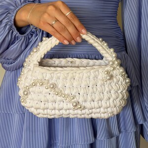 Puede incluir: Un bolso de mano blanco tejido a ganchillo con un asa trenzada y una decoración de perlas blancas de varios tamaños. El bolso tiene una forma redondeada y estructurada con un patrón de punto grueso y texturizado y un cierre metálico plateado.