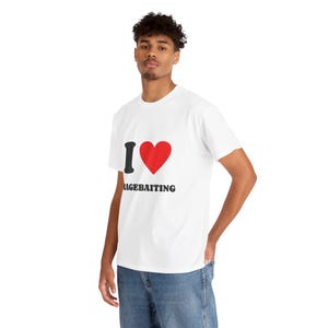 Op de afbeelding: Een wit T-shirt met korte mouwen en een grafische print op de borst. Het ontwerp bestaat uit een zwarte letter I, een rood hartsymbool en het woord RAGEBAITING in dikke zwarte hoofdletters onder het hart gedrukt.