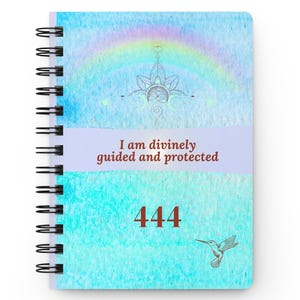 May include: A spiral-bound notebook with a light blue watercolor background featuring a faint rainbow, a central lotus-like line art symbol, and the text "I am divinely guided and protected" above the number "444" and a small hummingbird illustration.