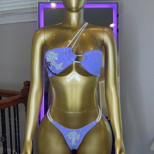 Peut inclure: Un ensemble bikini deux pièces lavande avec de fines bretelles blanches et une décoration en forme de croix sur le haut et le bas. Le tissu est orné de petits strass dispersés, et le haut présente un design de bretelle asymétrique unique.