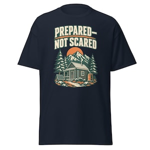 Può includere: Una t-shirt blu navy a maniche corte con una stampa grafica di una baita nel bosco con montagne e un sole sullo sfondo. Il testo PREPARED-NOT SCARED è stampato in color crema sopra la baita, che include un pannello solare e un barile d'acqua.
