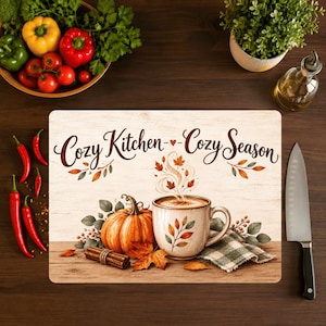 May include: A rectangular chopping board featuring a wood grain background with the text Cozy Kitchen Cozy Season, an illustration of a pumpkin, a mug with steam, cinnamon sticks, and a green plaid cloth, all decorated with autumn leaves.