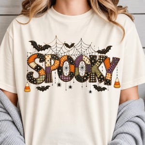 May include: A cream-colored t-shirt featuring the word SPOOKY in a patchwork-style font with various patterns including plaid, stars, and polka dots. The design includes black spiderwebs, hanging spiders, bats, and candy corn accents.