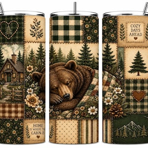 May include: A cylindrical tumbler with a straw featuring a patchwork design of a sleeping brown bear, a log cabin, pine trees, flowers, and plaid patterns. Text reads "HOME IS WHERE THE CABIN IS" and "COZY DAYS AHEAD" with heart and pine tree icons.