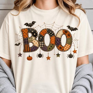 May include: A cream-colored t-shirt featuring a large graphic of the word BOO in a patchwork pattern of orange, black, and green. The design includes spiderwebs, hanging spiders, bats, stars, and pumpkins in a festive Halloween-themed style.