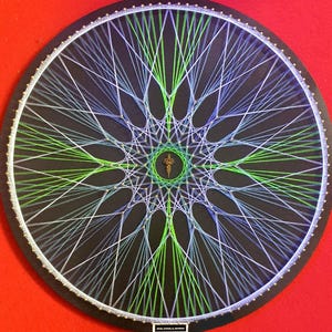 Puede incluir: Una pieza de arte de hilo redonda, con un fondo negro y un complejo patrón geométrico. El diseño utiliza hilos blancos, verdes y azules. La obra de arte está sobre un fondo rojo.