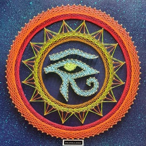 Puede incluir: Obra de arte en hilo que representa el Ojo de Horus. La obra utiliza hilos naranjas, amarillos y azules sobre un fondo azul oscuro. El diseño es circular con un patrón de estrella alrededor del ojo.