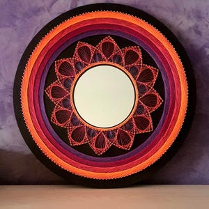 Puede incluir: Espejo de pared redondo negro con un diseño de hilo vibrante. El diseño presenta círculos concéntricos de hilo naranja, rojo y morado, que rodean un espejo central. El diámetro es de aproximadamente 30 cm.