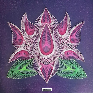 Puede incluir: Una pieza de arte de hilo que representa una flor de loto. La flor presenta hilos rosas y blancos para los pétalos, y hilos verdes para las hojas, todo sobre un fondo morado oscuro. El diseño es geométrico y simétrico, con una pequeña etiqueta rectangular en la parte inferior.