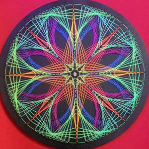 Könnte beinhalten: Ein kreisförmiges String-Art-Stück mit schwarzem Grund. Das Design zeigt ein symmetrisches Sternmuster, das mit leuchtenden neonfarbenen grünen, orangefarbenen, rosa und blauen Fäden erstellt wurde. Das Kunstwerk ist vor einem roten Hintergrund angeordnet.