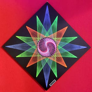 Puede incluir: Una pieza de arte de hilo geométrico sobre un cuadrado negro. El diseño presenta una espiral rosa central rodeada por un patrón de estrella en hilos verde neón, naranja y azul. La obra de arte está montada sobre un fondo rojo.