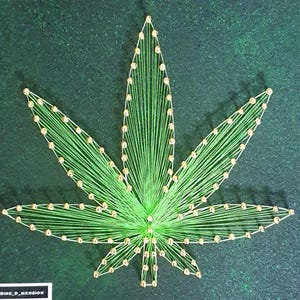 Può includere: Un design di arte con filo verde di una foglia di cannabis con accenti color oro. La foglia è centrata su uno sfondo verde scuro. L'arte con filo è realizzata con filo verde e spilli color oro.