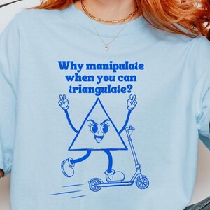 Op de afbeelding: Een lichtblauw T-shirt met korte mouwen en een donkerblauwe afbeelding van een cartoon-driehoek op een step. De tekst boven het figuur luidt: "Why manipulate when you can triangulate?". Het shirt heeft een ronde hals en een losse pasvorm.