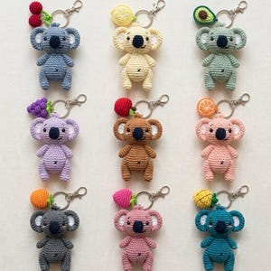 May include: A collection of nine crocheted koala keychains, each featuring a unique fruit or vegetable charm attached to a silver metal ring. The koalas are rendered in various colors including blue, yellow, green, purple, brown, pink, and teal.