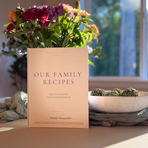 May include: A hardcover book with a cream-colored cover titled "OUR FAMILY RECIPES" in black serif font. Below the English title, the Armenian text "ՄԵՐ ԸՆՏԱՆԻՔԻ ԲԱՂԱԴՐԱՏՈՄՍԵՐԸ" is printed, followed by the author name "Սերգեյ Պայլյանին" at the bottom.