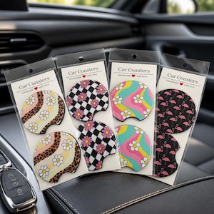 May include: Four sets of two circular car coasters in clear plastic packaging. Each set features distinct patterns: leopard print with daisies, black and white checkerboard with pink flowers, wavy rainbow stripes with smiley faces, and pink flamingos on black.