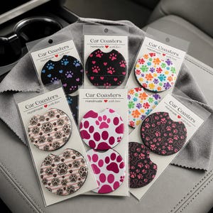 May include: Six clear plastic packages containing round car coasters with various designs including paw prints, bones, and hearts. Each package features a white header card with the text Car Coasters Handmade with love printed in black.