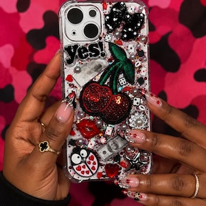 May include: Clear phone case decorated with cherries, dice, pearls, a bow, and the word "yes!". The case has a variety of red, black, and silver embellishments. The background is a pink camouflage pattern.