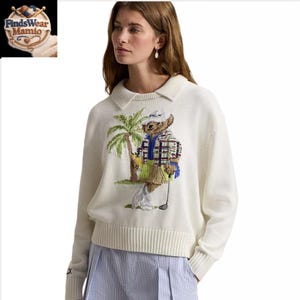 May include: Cream-coloured sweater with a collared neckline and a golf-themed embroidered design. The design features a bear wearing a plaid shirt and a golf hat, holding a golf club near a palm tree. The sweater has ribbed cuffs and hem.
