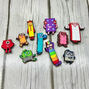 May include: A collection of ten colorful Numberblocks character charms made of rubber. The set includes various shapes and colors representing numbers, featuring smiling faces, limbs, and distinct patterns like dots, squares, and rainbow stripes.