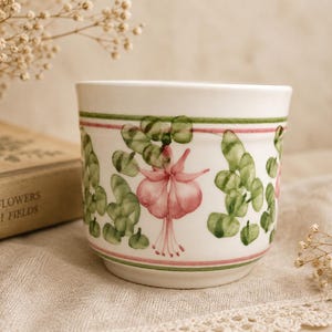 May include: A white ceramic planter featuring a hand-painted botanical pattern of pink fuchsia flowers and green foliage. The design is framed by thin horizontal pink and green lines encircling the top and bottom of the cylindrical vessel.