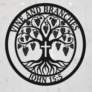 Könnte beinhalten: Ein kreisförmiges Wandkunstobjekt aus schwarzem Metall, das einen stilisierten Baum mit Weintrauben und einem zentralen Kreuz zeigt. Der Text VINE AND BRANCHES ist oben gebogen, und der Text JOHN 15:5 ist unten am kreisförmigen Rahmen gebogen.