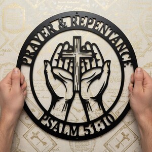 Könnte beinhalten: Ein kreisförmiges Wandkunstobjekt aus schwarzem Metall mit einem ausgeschnittenen Design von zwei gefalteten Händen, die ein Kreuz halten. Der Text PRAYER & REPENTANCE ist oben und PSALM 51:10 unten im kreisförmigen Rahmen eingearbeitet.