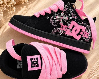 May include: A pair of black and pink DC sneakers featuring thick pink laces, a black suede upper with white floral-inspired patterns, a pink DC logo on the side and tongue, and a silver cross charm attached to the laces near the eyelets.