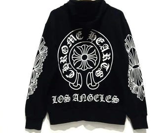 Könnte beinhalten: Ein schwarzer Kapuzenpullover mit einer großen weißen kreisförmigen Grafik auf der Rückseite mit dem Text Chrome Hearts und Los Angeles. Die Ärmel sind mit weißen floralen Kreuzmustern verziert und die Kapuze ist am Halsausschnitt befestigt.