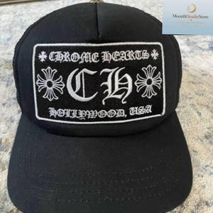 May include: A black baseball cap featuring a rectangular patch on the front. The patch displays the white embroidered text CHROME HEARTS, the letters CH, and the text HOLLYWOOD, USA, flanked by two white cross symbols on a black background.