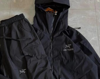 May include: A black hooded jacket and matching cargo trousers set. The jacket features a full-length front zip, a hood, and the white Arc'teryx logo on the chest. The trousers include side cargo pockets and the white Arc'teryx logo on the leg.