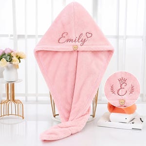 Pode incluir: Uma toalha de cabelo com capuz em rosa suave, apresentando o nome Emily bordado, um pequeno ícone de coração e um fecho de botão em forma de urso. Um círculo inserido mostra um design de bordado alternativo com uma coroa sobre a letra E.