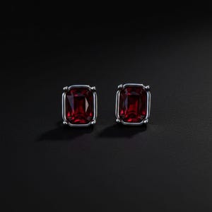 Puede incluir: Un par de pendientes de botón con piedras preciosas rectangulares de color rojo intenso engastadas en metal plateado. Los pendientes se presentan sobre un fondo oscuro, resaltando el color vibrante y la forma de las piedras. El diseño es simple pero elegante.