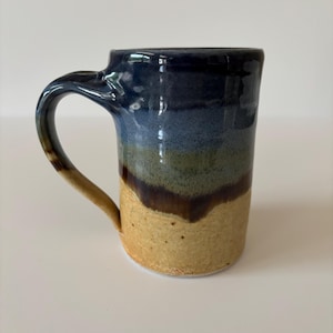 May include: A ceramic mug featuring a vertical gradient of colors, transitioning from a dark blue rim down to a muted green middle section, and ending in a textured, sandy beige base. The mug has a curved handle and a smooth, glossy finish.