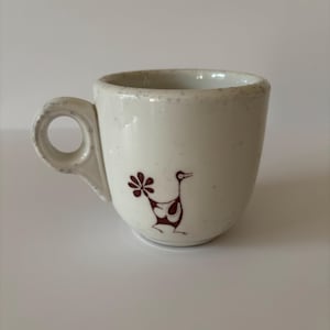 May include: A cream-colored ceramic mug with a speckled finish and a circular handle. The mug features a small, dark brown graphic of a stylized bird with a fan-shaped tail on the front surface. The design is simple, featuring a rounded body.