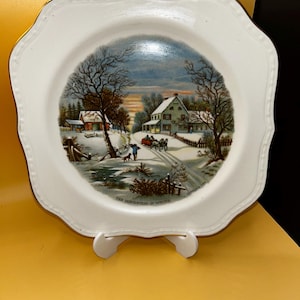 May include: A white ceramic plate with a scalloped edge and gold trim featuring a Currier and Ives winter scene titled The Homestead in Winter. The illustration depicts a snow-covered landscape with a house, a horse-drawn carriage, and trees.
